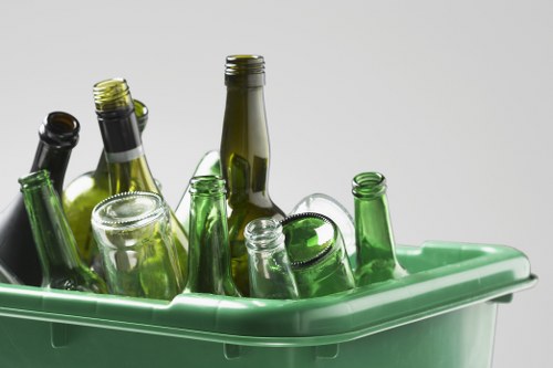 A green plastic recycling bin filled with empty glass bottles of various sizes and colors, predominately green and clear, with some dark brown bottles, all placed on the edge of a plain, light grey background. The bottles are upright or angled, with some appearing slightly tilted or stacked against each other, showing smooth glass surfaces and reflective finishes. The bin is positioned outdoors, possibly on a driveway or pavement, with no other objects visible in the immediate surroundings. The scene emphasizes the collection of recyclable glass waste, consistent with rubbish removal services offered by Commercial Waste Highbury, situated within the Highbury area. The natural lighting highlights the transparency and textures of the glass bottles, reinforcing environmental and recycling themes integrated into local waste management practices.